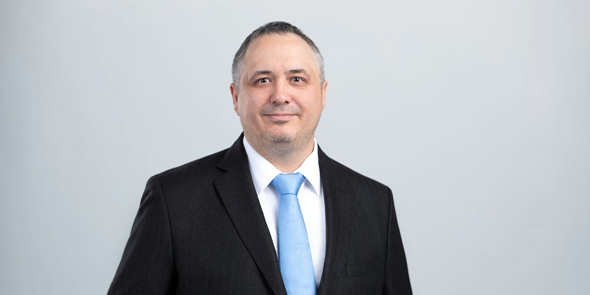 Ein Mann mit kurzem dunklem Haar lächelt leicht und trägt einen schwarzen Anzug mit hellblauer Krawatte